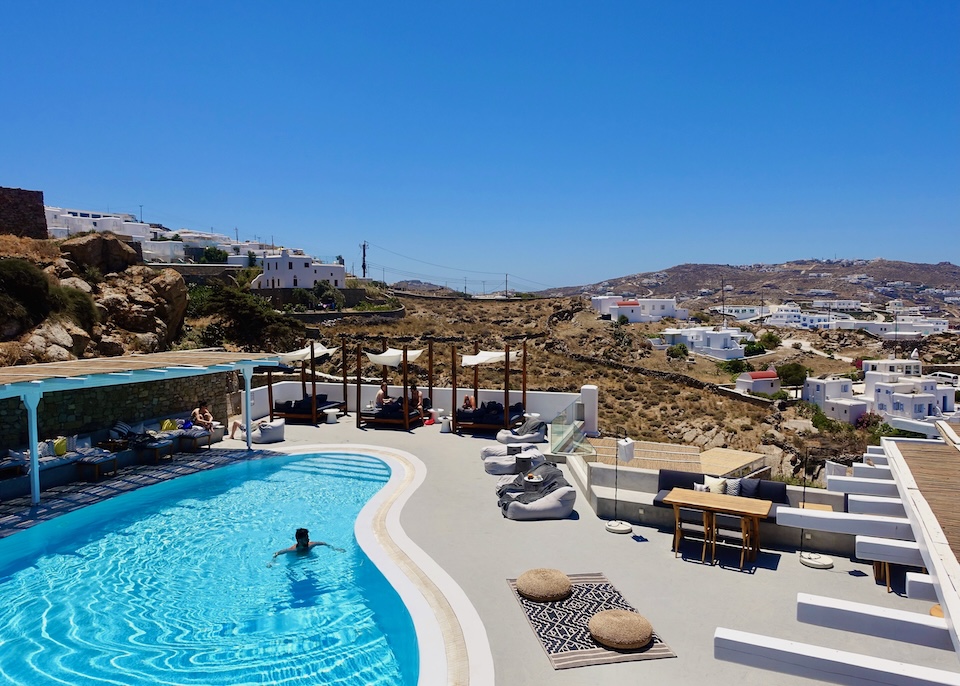 A kidney-shaped pool and deck, partially shaded by a pergola, facing the town and hills at Boheme hotel on the edge of Mykonos Town.