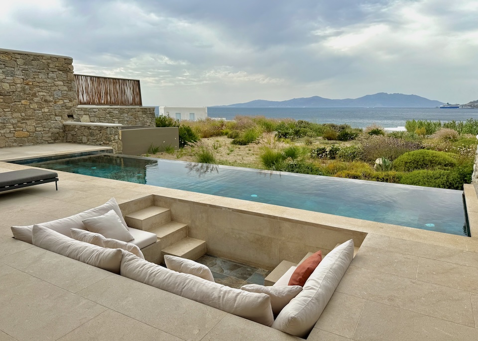 A sunken sofa and lounge space behind an infinity pool facing a garden and the sea at Bonzoe Suites and Villas in Megali Ammos, Mykonos.