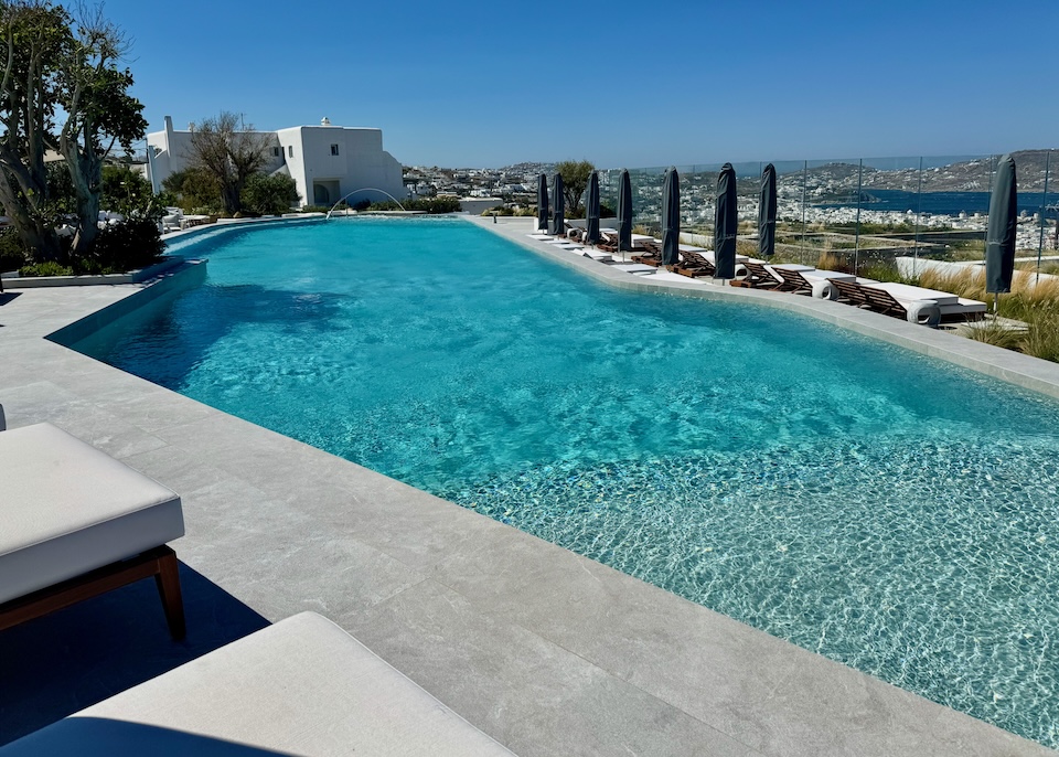 A freeform pool with sunbeds on two sides faces the sea and Mykonos Town at Deos hotel in Mykonos.