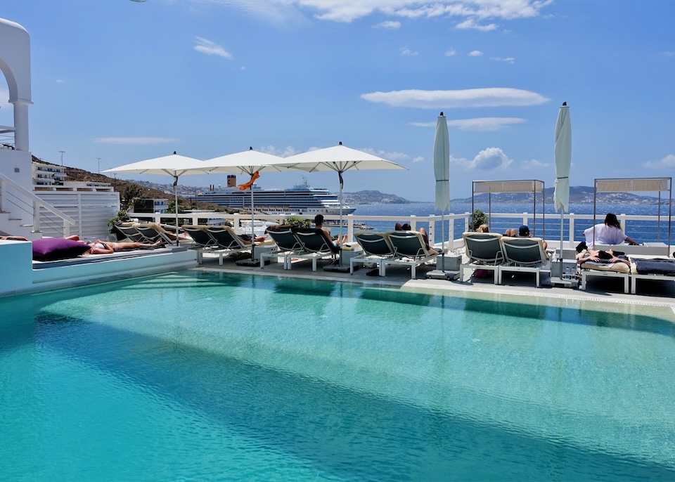 A swimming pool with a row of sunbeds in pairs all facing the sea and ferry port at Grace Hotel in Agios Stefanos, Mykonos.