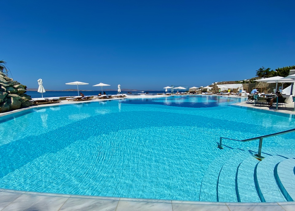 Steps lead down into a large freeform infinity pool with sunbeds at the far end overlooking the sea at Mykonos Grand resort in Agios Ioannis, Mykonos.