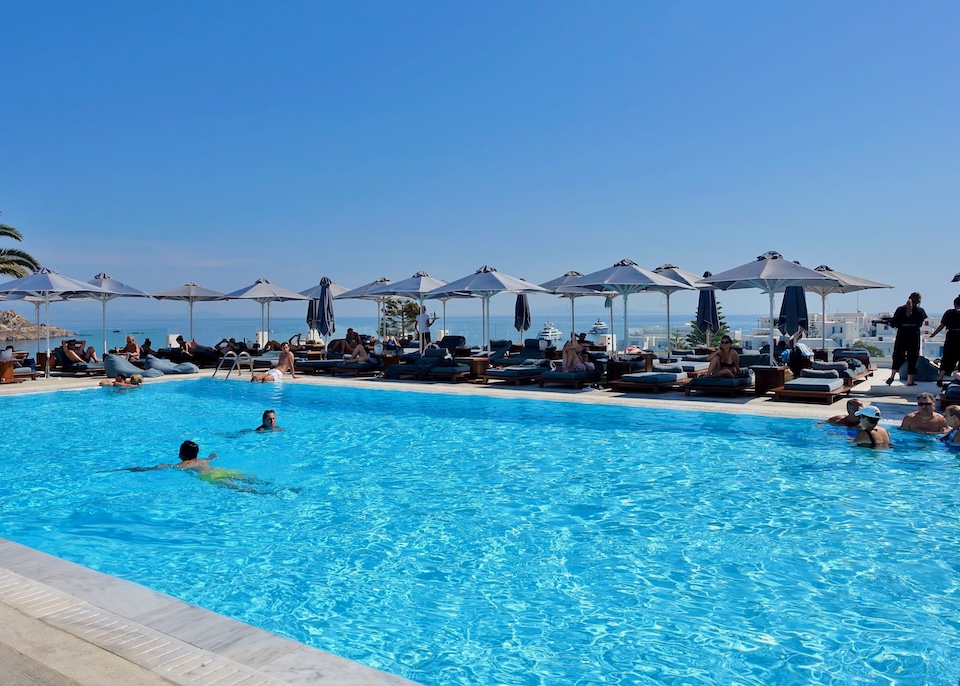 Swimmers in a rectangular pool with sunbeds and umbrellas on the far side and the sea in the distance at Myconian Ambassador hotel in Platis Gialos, Mykonos.