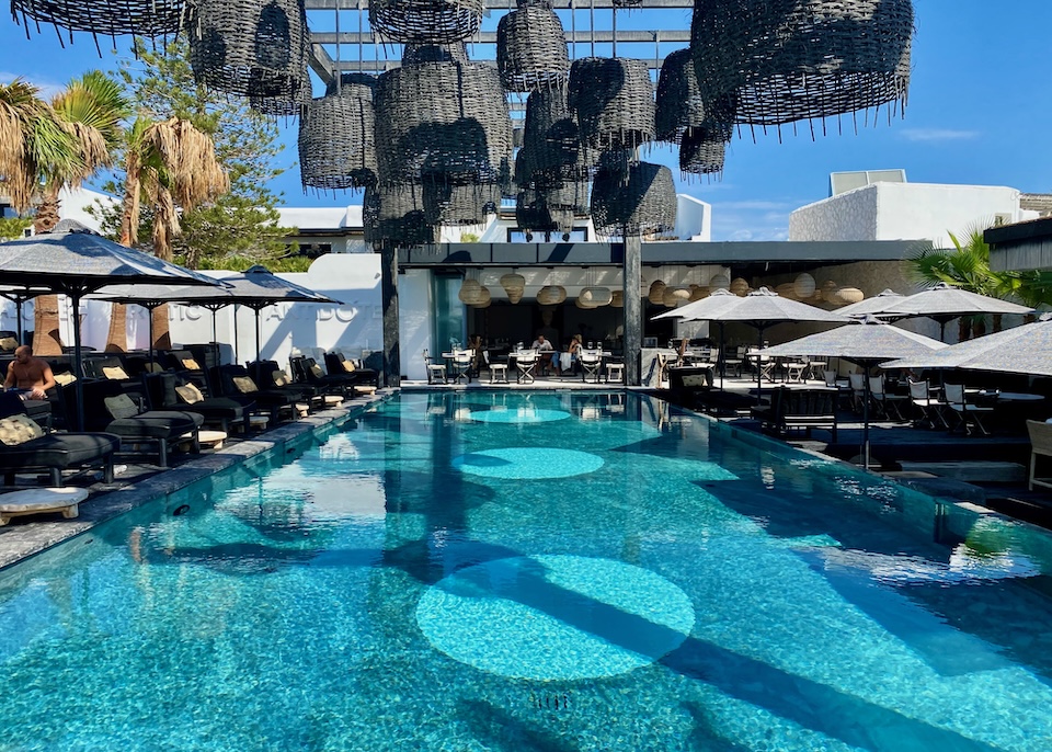 A rectagular infinity pool sits under basket-style pendant lights with sunbeds on the sides and a restaurant in the back at Myconian O hotel in Ornos, Mykonos.