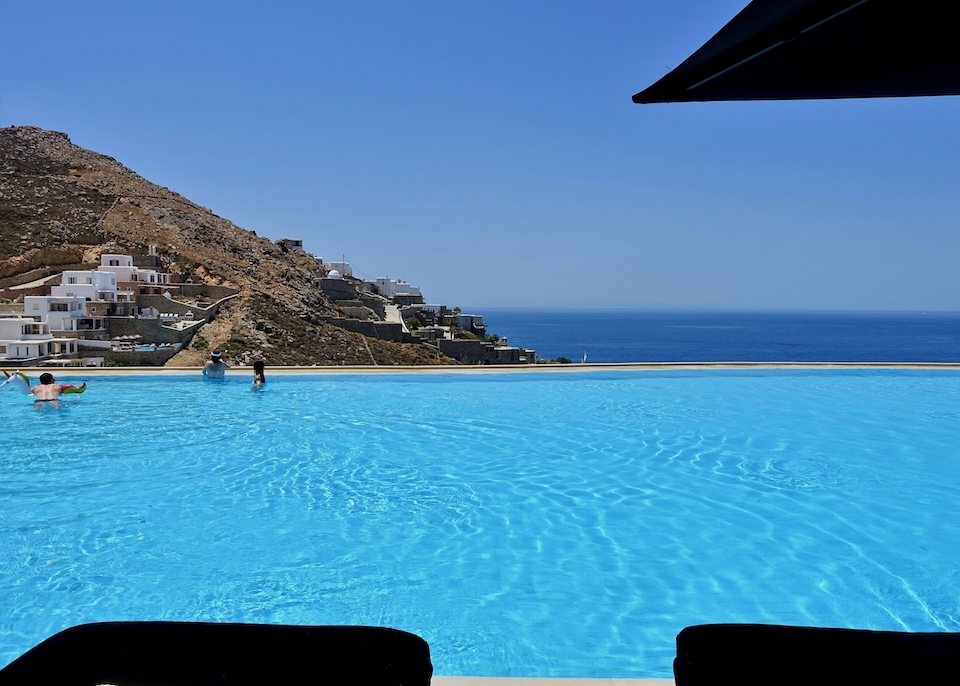 People swimming in an infinity pool that overlooks the sea and hills at Myconian Villa Collection in Elia, Mykonos.