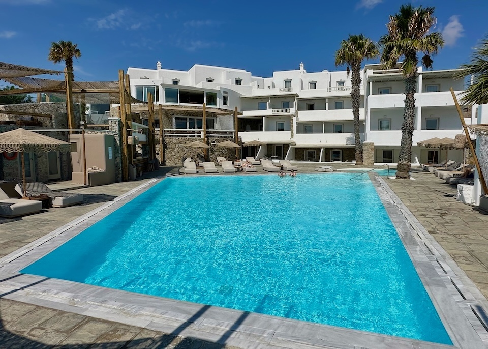 An angular infinity pool in front of a whitewashed, four-story hotel and palm trees at Palladium Boutique Hotel in Platis Gialos, Mykonos.