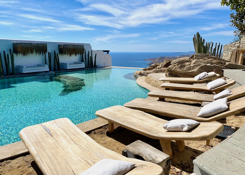 A row of wooden sunbeds lines a freeform infinity pool above the sea at Panoptis Escape resort in Elia, Mykonos.