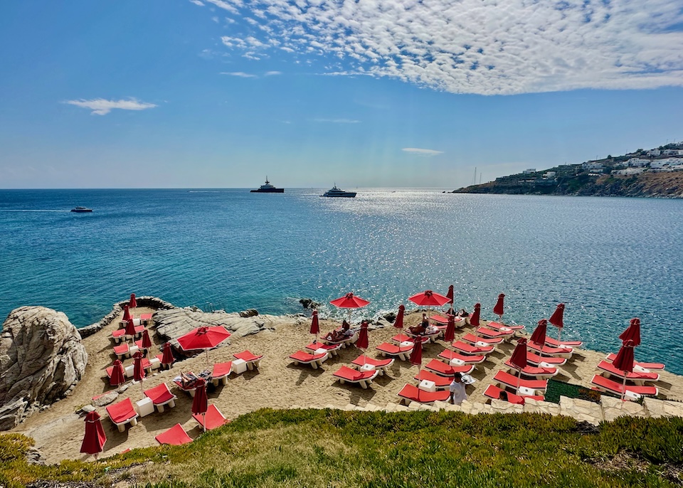 A cluster of red sunbed and umbrella sets sit on a private beach with a view to the sea and boats at Petasos Beach Resort in Platis Gialos, Mykonos.