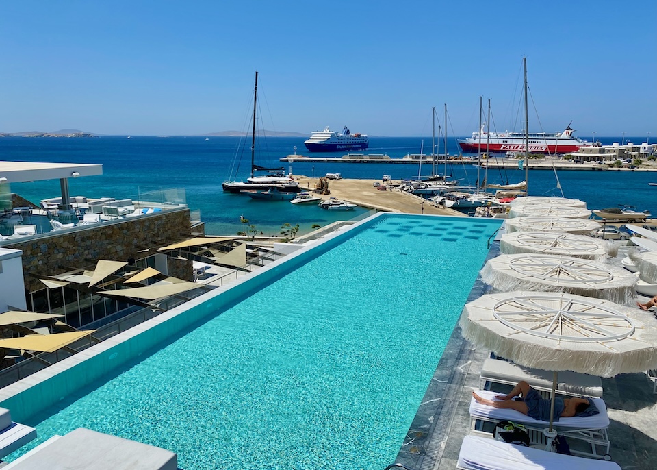 An infinity pool lined with sunbeds and umbrellas stretches out toward the sea with a view of the ferry port and marina at Mykonos Riviera in Tourlos, Mykonos.