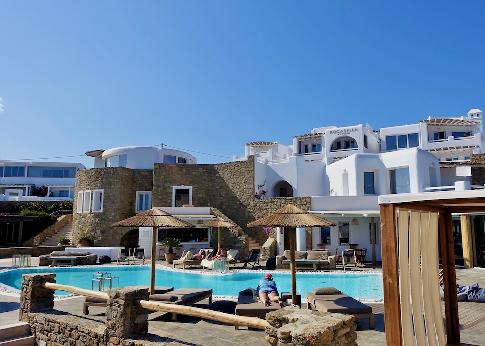 A pool sits in front of three story building, half of it made with natural stones and half of it whitewashed at Rocabella hotel in Agios Stefanos, Mykonos.