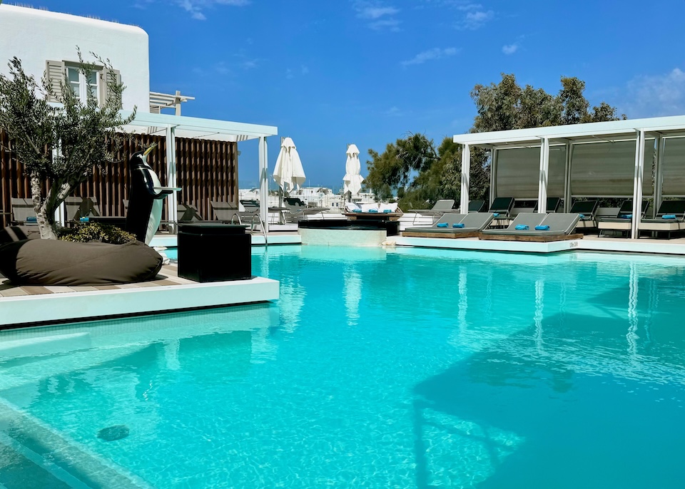 A central pool with sunbeds and pergolas around and a penguin statue next to an olive tree at Semeli Hotel in Mykonos Town.