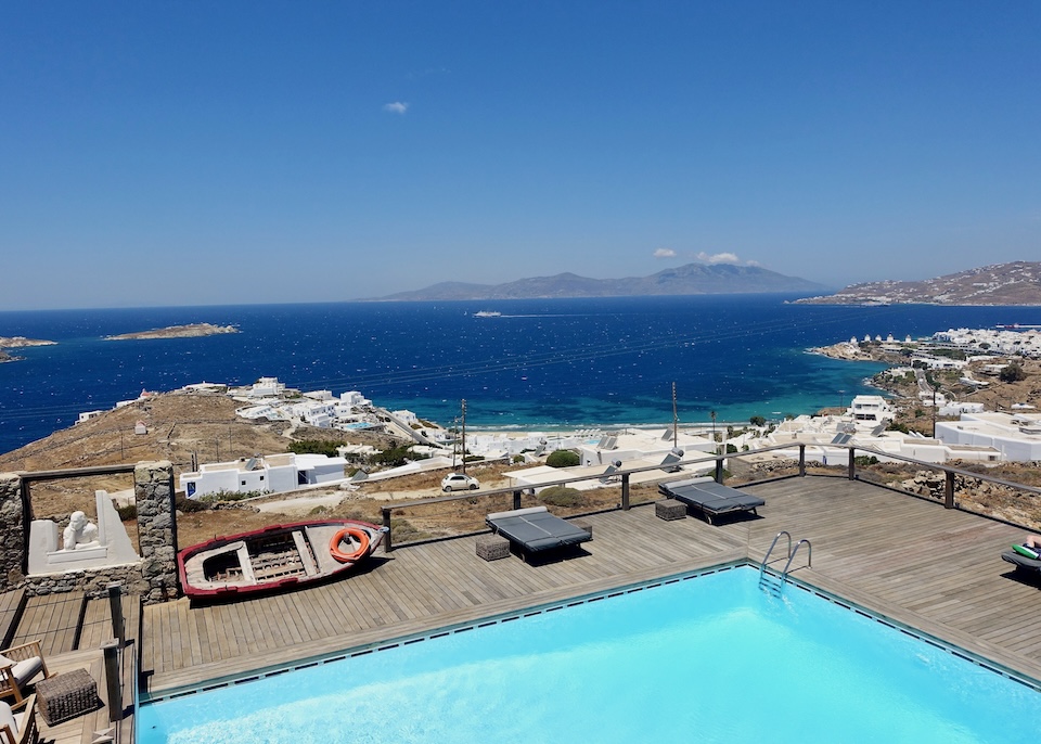 A square pool with a wooden sun deck overlooking the sea toward Mykonos Town at Tharroe of Mykonos hotel.