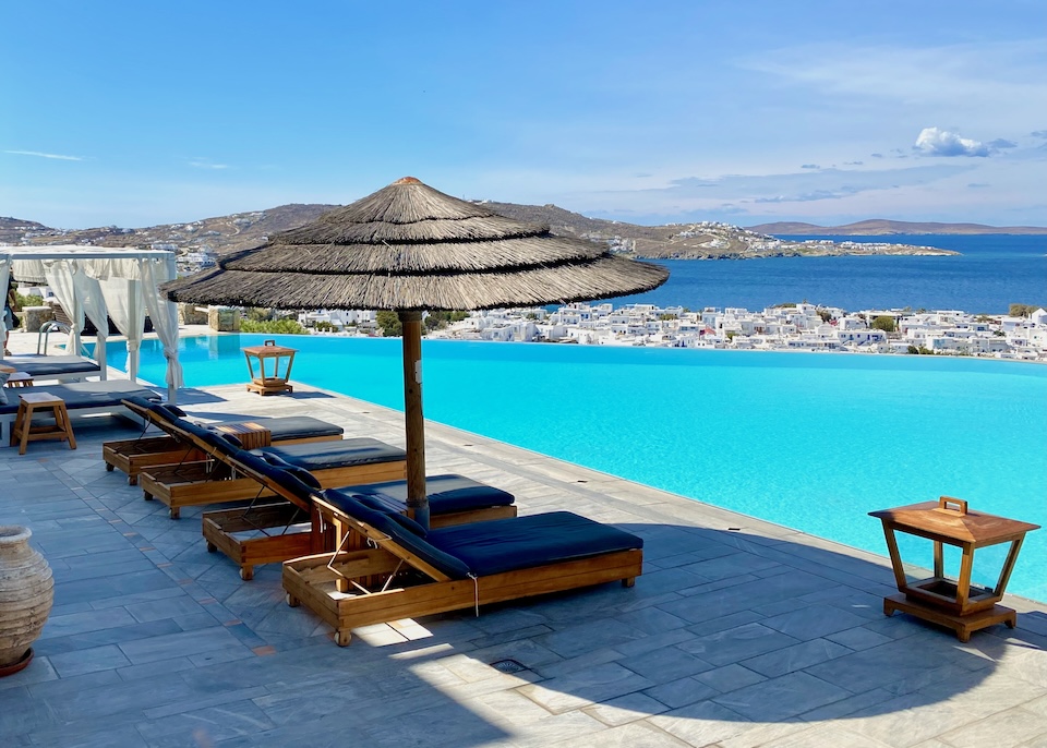 One thatched umbrella at the start of a row of sunbeds and Bali beds, all sitting behind an infinity pool overlooking Mykonos Town and the sea at Vencia Boutique Hotel.