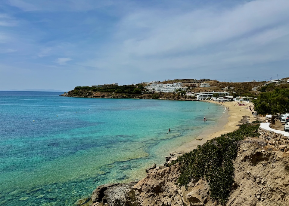 View from above crescent-shaped Agios Stefanos Beach with varied hues of turquoise water in Mykonos.