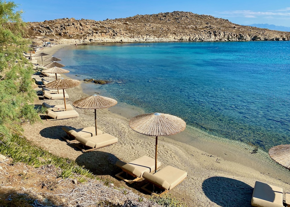 Pairs of sunbeds, each with a thatched umbrella, on a sandy beach with no waves in Aleomandra, Mykonos.