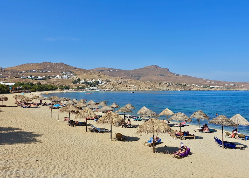 A wide, sandy beach with thatched umbrellas, sunbeds, and people lounging at Kalafatis Beach in Mykonos.