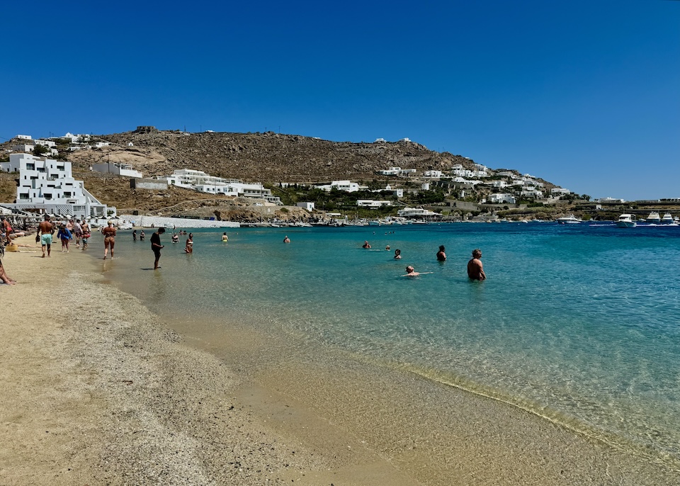 Beachgoers swimming and strolling along Ornos Beach in Mykonos.