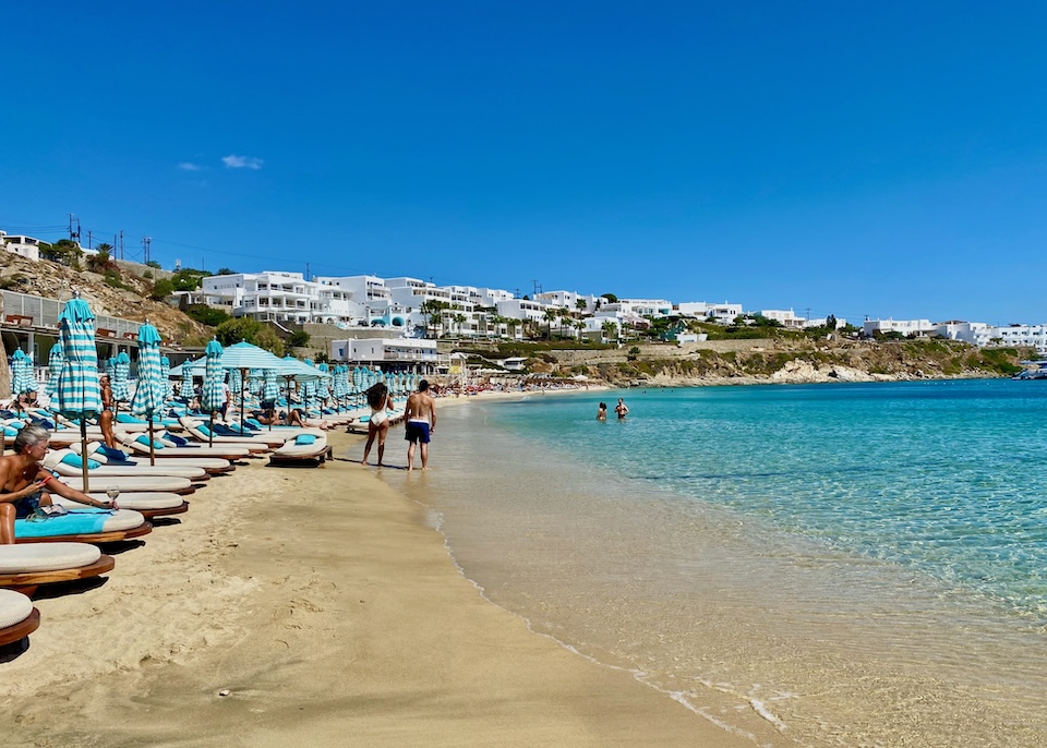 A few people walking, lounging, and swimming at Psarou Beach in Mykonos.