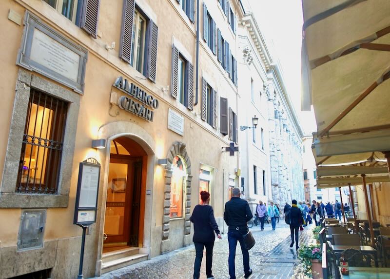 Pedestrians walking down a street in Rome past a hotel building and cafe tables