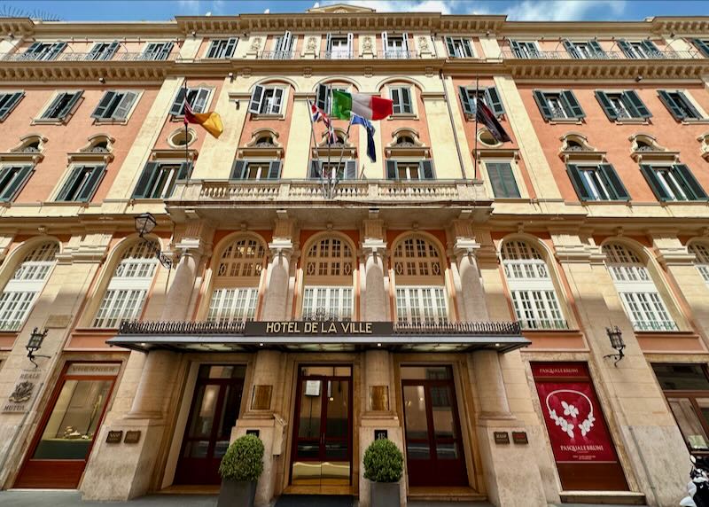 View looking up at an ornate peach-colored hotel in Rome, festooned with flags