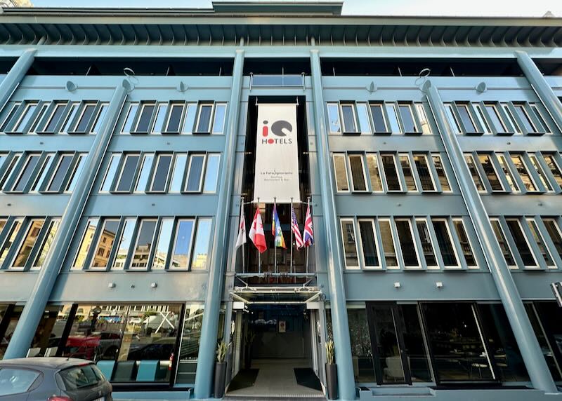 Exterior of a large modern blue hotel building with tall windows and flags flying above the doorway