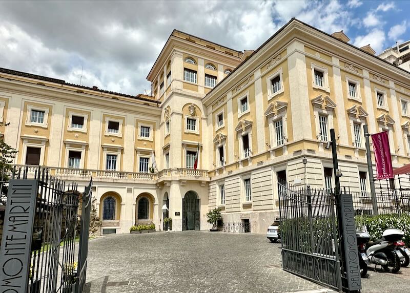 Open wrought-iron gates leading to the drive of a stately cream-colored Italian hotel 