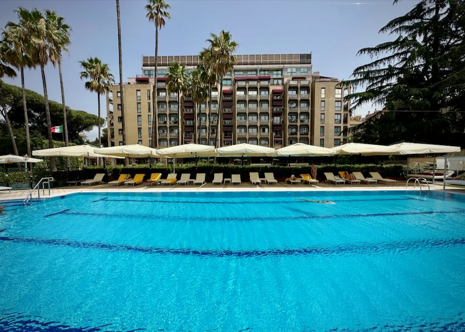 View across a large swimming pool to a hotel flanked by palm trees