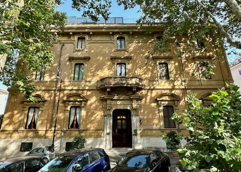 Sun-dappled exterior of a yellow classic hotel building in Rome, with cars parked in front