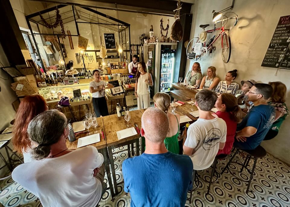 Two people present food options to a crowd of onlookers in a rustic Italian bar.