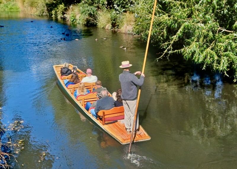 A man uses a long stick to row a flat boat of people through a river.