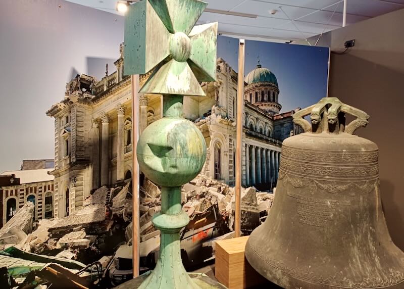 A large light green cross with a dent in it sits next to a large steel bell.