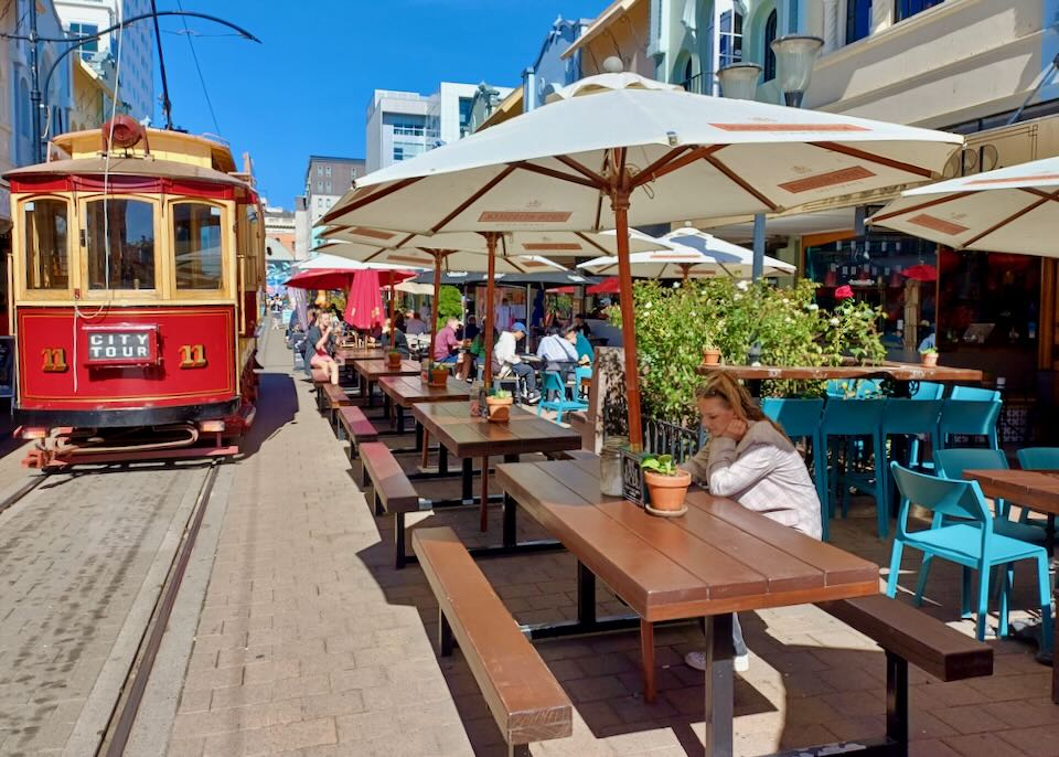 A red electric trolly rolls past wooden picnic tables on a brick street.