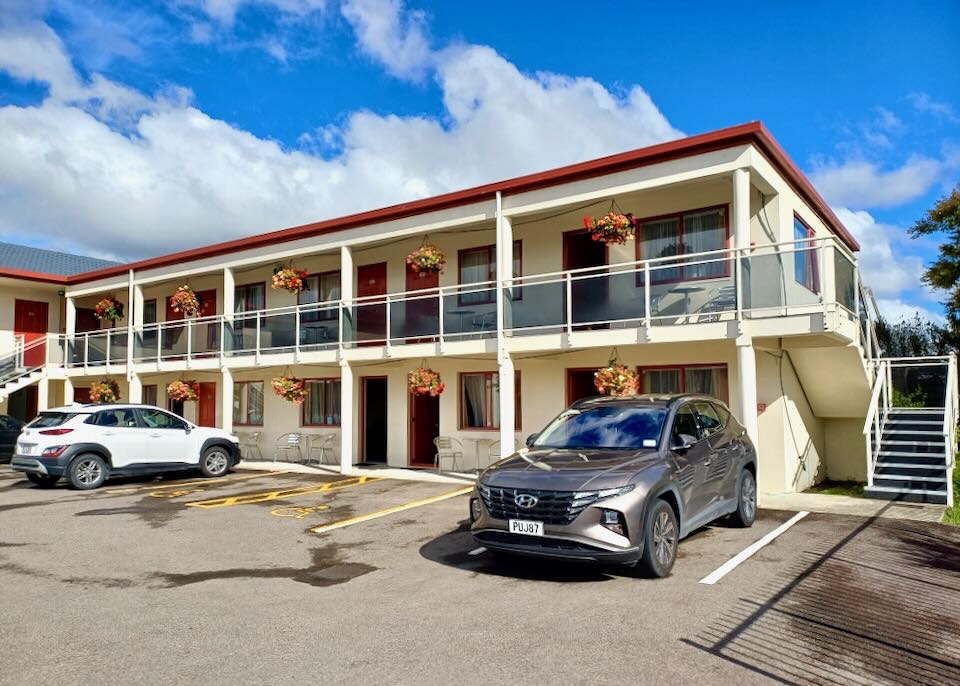 A long cream colored motel with red trim around the windows has hanging flower baskets on the balcony.