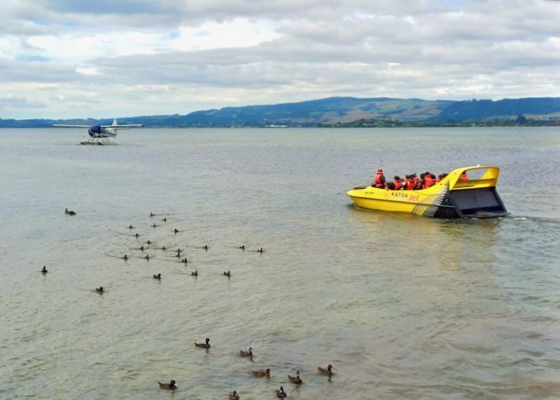 A bright yellow boat with passengers wearing red life jackets waits for a seaplane on the lake.