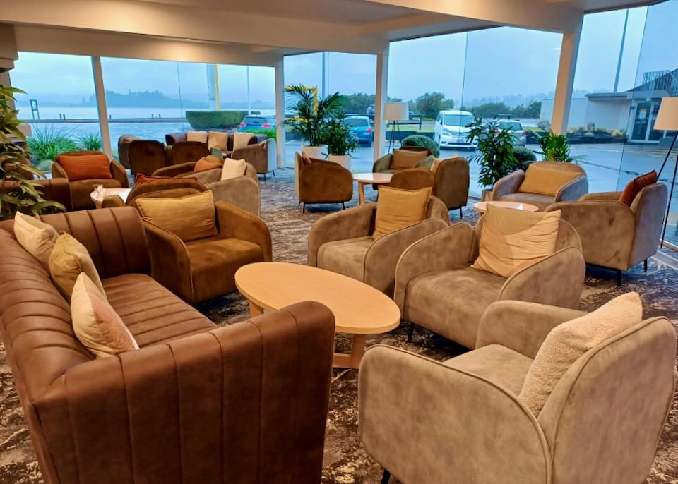 Large brown chairs fill a hotel lobby with walls of windows facing a lake.