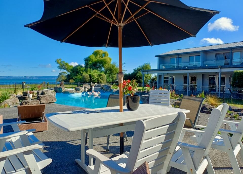 A table with a black umbrella sits next to a blue pool and in the distance is a lake.