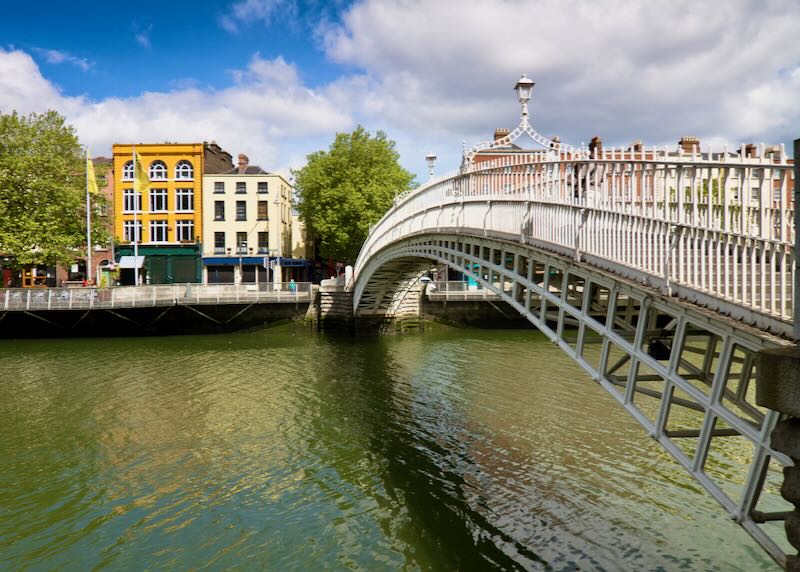 White pedestrian bridge across a greenish river, with colorful buildings on the opposite side.