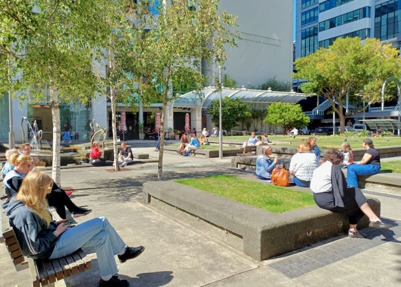 People sit on benches at a green park near a tall building.