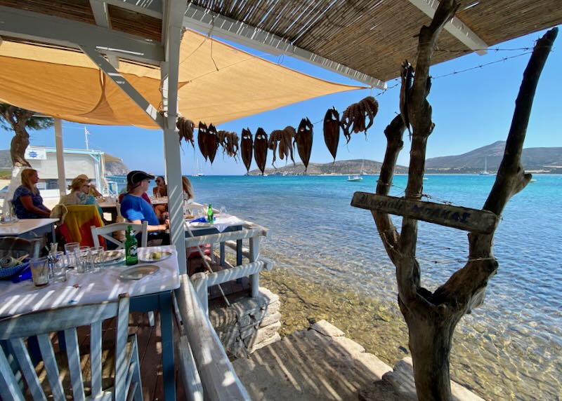 People sit at a dining balcony overlooking the water, while fish dry on a line in the sun next to them