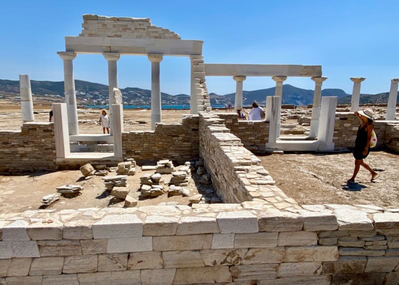 Tourists walk around ruins of an ancient temple with the sea in the background.