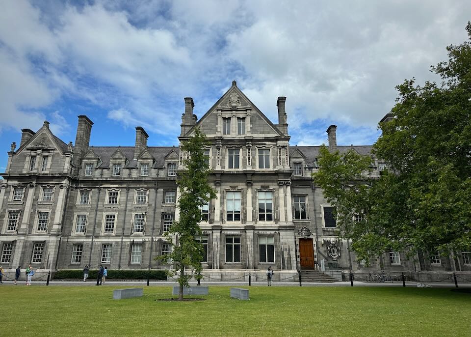 Front view across a green lawn of the grey stone buildings of Trinity College in Dublin on a sunny day.