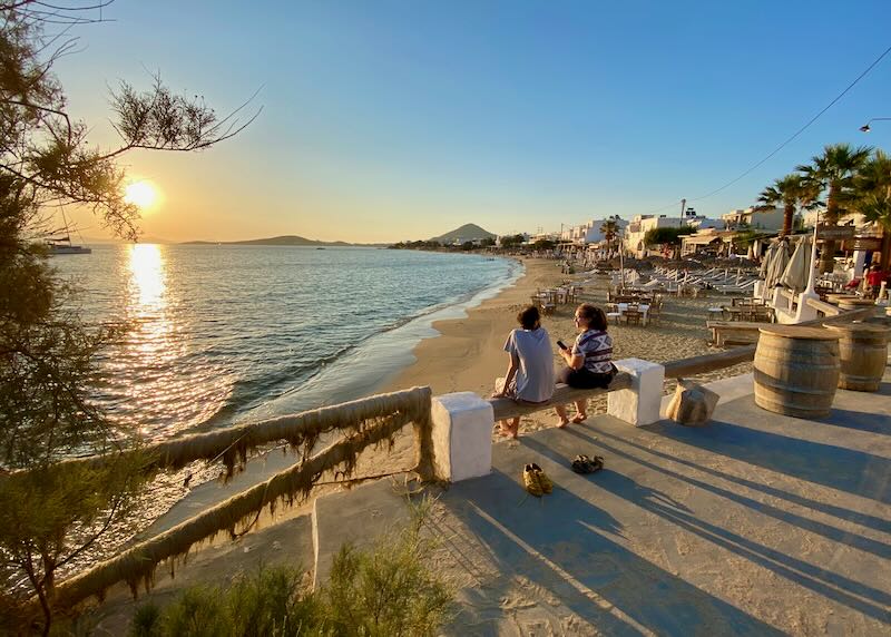 Two women sit and watch the sun set at a beach lined with shops and restaurants.