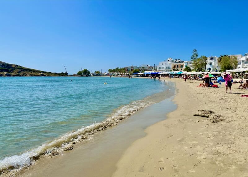 View looking up a golden sandy beach toward a town with boxy white Greek buildings.