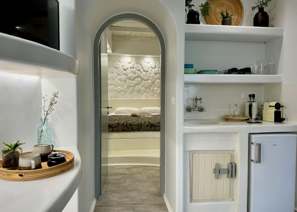 View looking past a kitchenette through an open arched doorway to a hotel bed in Cycladic style.