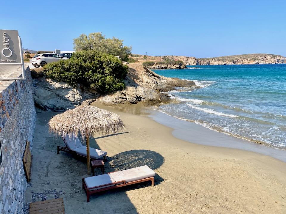 Sun beds shaded by thatched umbrellas on a sandy beach in a rocky cove. 