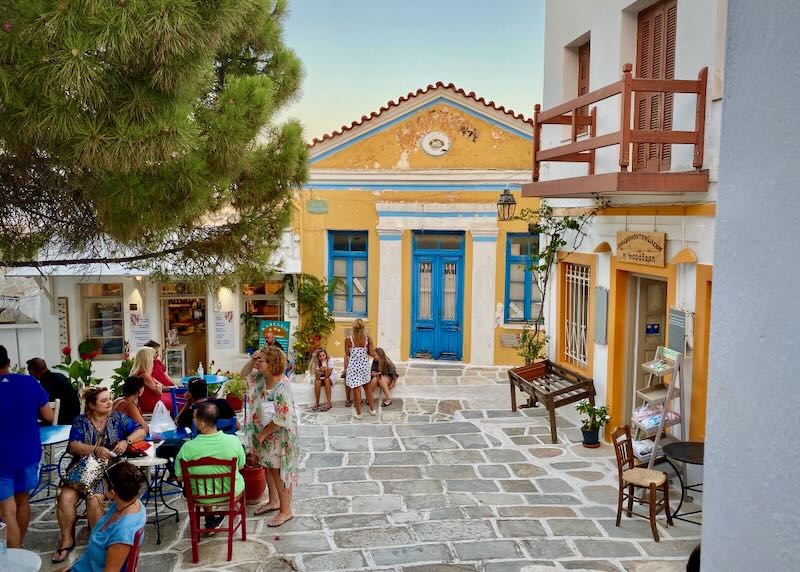 People sitting at small tables at a cobbled plaza in a Greek village