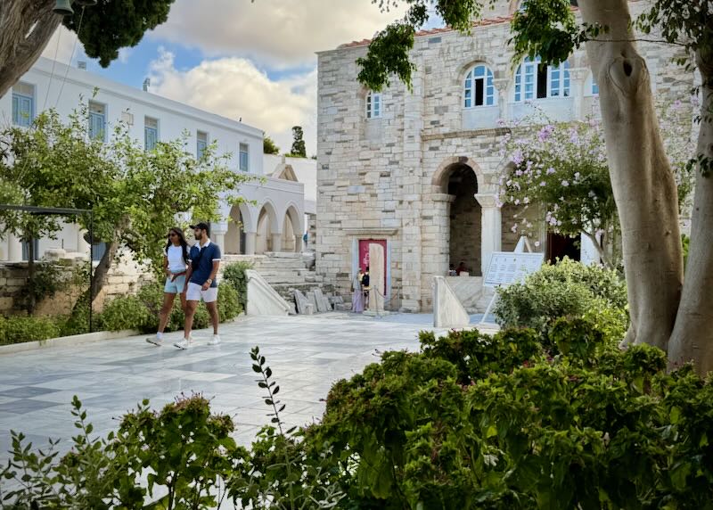 Two people walk in a cobblestone courtyard of a stone church.