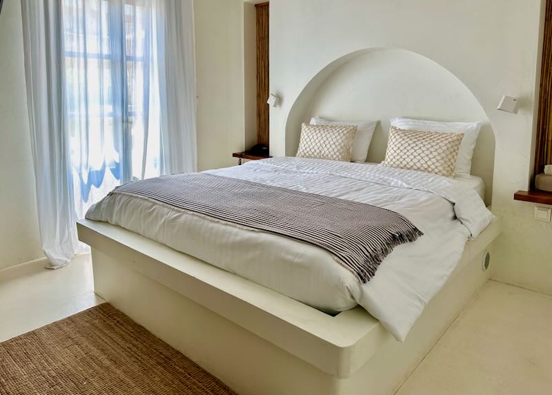 Inviting looking hotel bed with white puffy covers, next to a balcony door.