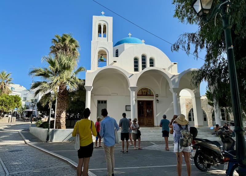 Tourists take photos of a blue-domed white church in a Greek village.