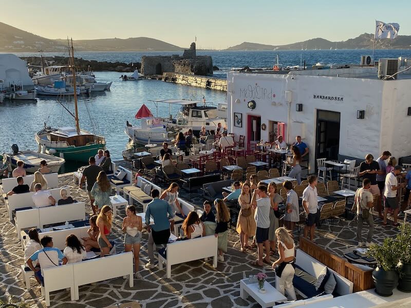 People sit at restaurant tables at a Greek harbor at sunset.