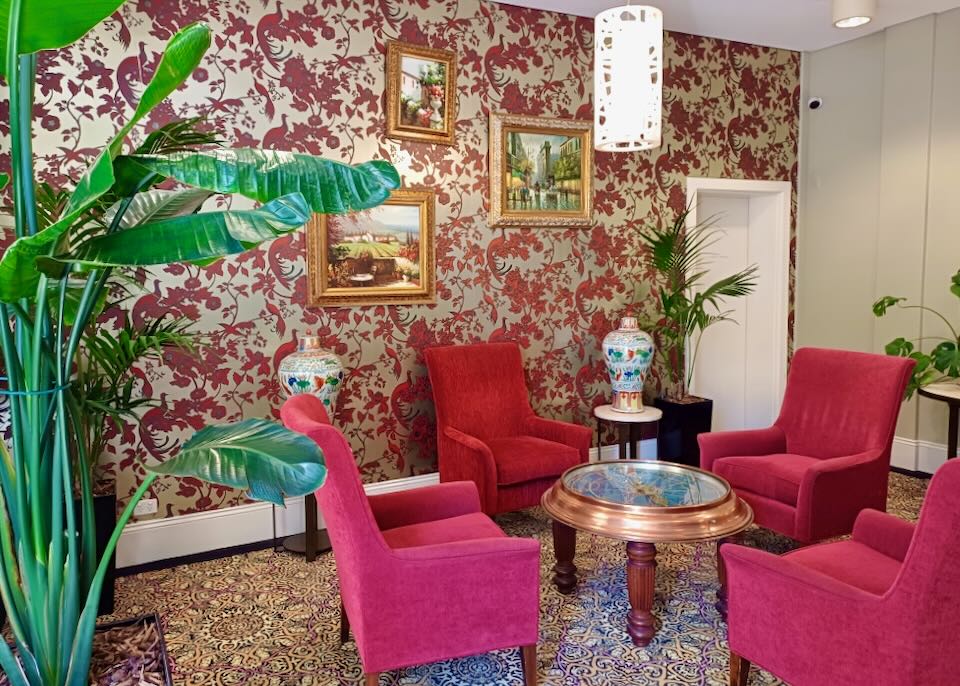 Bright pink chairs sit in a hotel lobby with pink and gold floral wallpaper and large green plants.